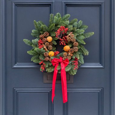 Çam, kozalak, tarçın çubukları, kurutulmuş portakal dilimleri ve kırmızı böğürtlenlerden oluşan geleneksel bir Noel çelengi koyu mavi bir kapı üzerinde parlak kırmızı ipek bir yayla asılır..