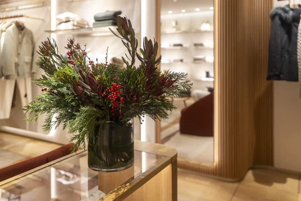 Yeşil çam yaprakları, kırmızı böğürtlenler ve koyu kırmızı çiçeklerden oluşan şenlikli bir düzenleme parlak ışıklandırılmış modern lüks giyim mağazasının içindeki cam bir vazoda duruyor..