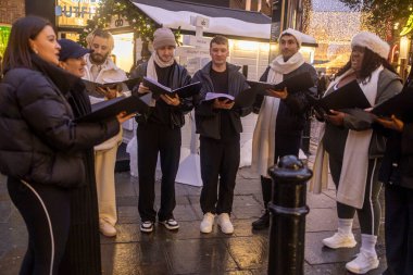Londra, İngiltere, 13 Aralık 2025. Bir grup genç Noel şarkıcısı, bir şehir sokağında kış kıyafetleri içinde şarkı söylüyor.