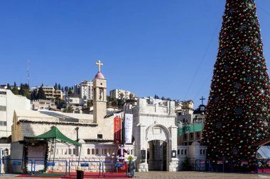 Nasıra, İsrail, 24 Aralık 2025, Yunan Ortodoks Annunciation Kilisesi. Taş binalar ve çan kulesi olan bir kilise meydanının yakınındaki büyük süslü Noel ağacı.