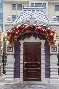 Moskova, Rusya - 30 Aralık 2025: Ahşap kapılı küçük bir bina ve kırmızı Noel mücevherleri ve karda çelenklerle süslenmiş kemer,