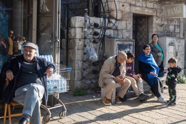 Nasıra, İsrail, 25 Aralık 2025. İnsanlar güneşli bir sokakta oturmuş, taş bir binanın yanında duruyor ve eski bir kasaba bölgesindeki günlük yaşamı gösteriyor.