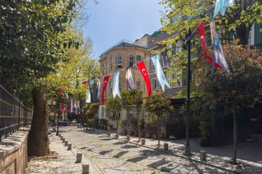 İstanbul, Türkiye, 23 Mayıs 2025, Sunny kaldırım taşı caddesi ağaçlarla ve asılı bayraklarla çevrili, sakin bir mahalle manzarası gösteriyor. Cihangir ilçesi