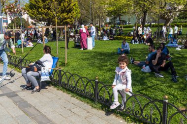 İstanbul, Türkiye, 5 Mayıs 2025, çeşitli kalabalıkların yeşil çimenler ve banklarda dinlendiği kamu parkı sahnesi. Ön planda siyah metal çitler,