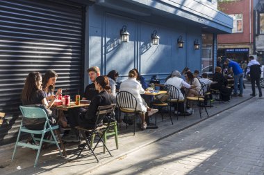 İstanbul, Türkiye, 5 Mayıs 2025, öğleden sonra saatlerinde yemek yiyen ve konuşan insanlarla dolu mavi duvar boyunca uzun açık hava kafe masaları. Ön planda bir grup genç arkadaş meşrubat içiyor.