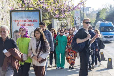 İstanbul, Türkiye, 5 Mayıs 2025, bazıları türban takmış erkekler ve kadınlar kiraz ağaçları ve kayalık yamaçlı geniş bir afişin önünden geçerek güneşli bir kaldırımda yürüyorlar. Uskudar 'daki sokak sahnesi, canlı bir yayaydı.