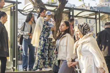 İstanbul, Türkiye, 5 Mayıs 2025 Çiçekli, uzun yeşil elbiseli genç bir kadın kucağında küçük bir çocukla büyük bir karganın içinde hareketsiz duruyor