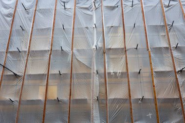 Kış boyunca üzerinde dikey ahşap kirişler ve beyaz koruyucu ağlar bulunan çok katlı bina cephesini kaplayan bir inşaat iskelesinin yakın çekimi. Kar, alt kenarların tozunu alır ve ince gölgeler, dalgalar boyunca geometrik desenler yaratır.