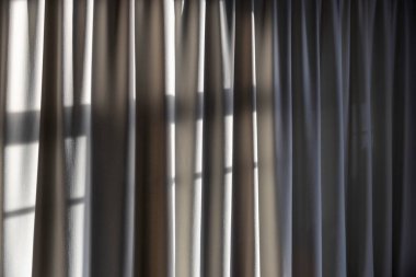 Katıksız keten ya da şeffaf kumaş perdelerin yakın çekim görüntüsü güçlü pencere ışığı ile tekstil yüzeyinde dramatik çizgili gölge desenleri yaratarak doku ve hareketi gösterir.