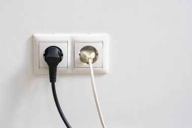 Biri siyah diğeri beyaz olmak üzere iki tane elektrik prizinin yakın plan görüntüsü düz bir duvardaki çift beyaz bir prize yerleştirilmiş.