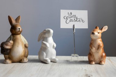 Tahta bir masanın üzerinde beyaz kart tutacağı olan üç farklı Paskalya tavşanı figürü (kahverengi, beyaz ve seramik). Kart Mutlu Doğular
