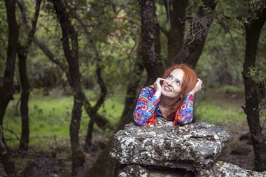 Kızıl saçlı genç bir kadın büyük bir taşa yaslanıyor ve yeşil bir ormana bakıyor..