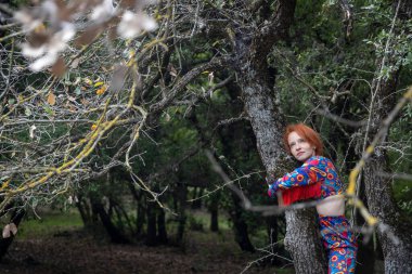 Kızıl saçlı hayalperest kadın çalılıklarda bir ağaca yaslanırken yukarı bakıyor..