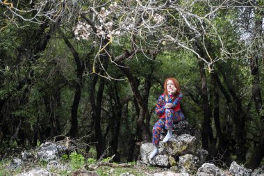 Vahşi bir ormanda, eski taşların üzerinde dinlenmiş canlı kıyafetli, şık kızıl saçlı bir kadın.
