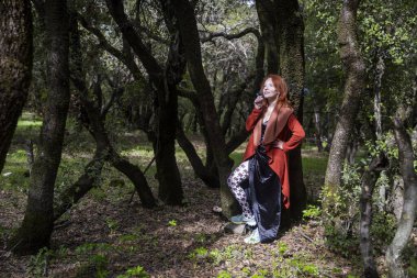 Kızıl saçlı, uzun kırmızı hırkalı ve kadife elbiseli bir kadın ormanda yaşlı ağaçların arasında poz veriyor.