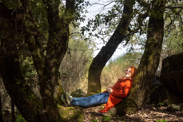 Turuncu kazaklı güzel kızıl saçlı kadın büyük yosunlu ağaç gövdesinde dinleniyor.