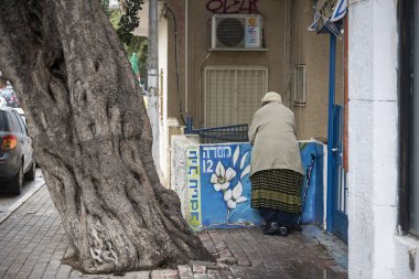 Hayfa, İsrail, 15 Şubat 2026 Masada caddesinde büyük bir ağaç gövdesinin yanında renkli bir duvar resminin yanında duran yaşlı bir kadının arka görüntüsü.