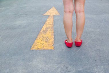 Kırmızı ayakkabılı bir kız asfalt bir arka planda sarı trafik okuna doğru yürüyor.