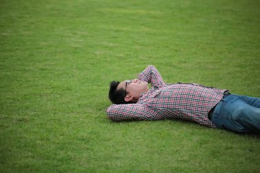 Yeşil çimenlerin üzerinde uzanan bir adam çek gömlek giyiyor, parkta kot pantolon giyiyor ve yaz günü gökyüzüne bakıyor.