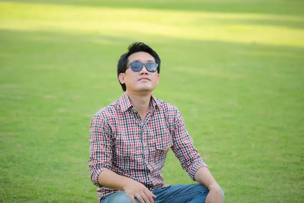 穿着格子衬衫 蓝色牛仔裤 戴着黑色太阳镜 坐在绿色草坪上的男人 — 图库照片