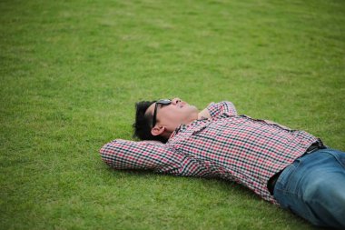 Yeşil çimenlerin üzerinde uzanan bir adam çek gömlek giyiyor, parkta kot pantolon giyiyor ve yaz günü gökyüzüne bakıyor.