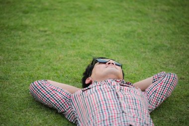 Yeşil çimenlerin üzerinde uzanan bir adam çek gömlek giyiyor, parkta kot pantolon giyiyor ve yaz günü gökyüzüne bakıyor.