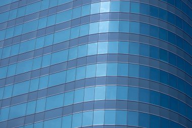 Modern bir binanın birçok dikdörtgen pencereli bir bölümü berrak, parlak mavi gökyüzünü yansıtıyor..