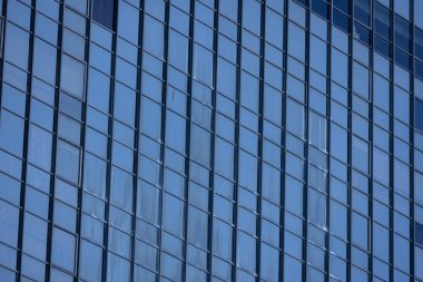 Modern bir binanın birçok dikdörtgen pencereli bir bölümü berrak, parlak mavi gökyüzünü yansıtıyor..