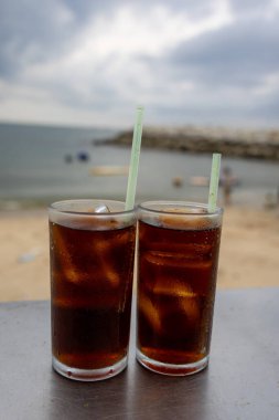 Bir bardak buzlu kola, deniz kenarındaki bir masada, bulutlu bir arka planda duruyor. İçkinin kabarcıkları ve yoğunlaşması, canlandırıcı kalitesini vurguluyor. Plaj manzarası rahatlatıcı bir yaz havası yaratıyor..