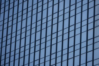 Modern bir binanın birçok dikdörtgen pencereli bir bölümü berrak, parlak mavi gökyüzünü yansıtıyor..