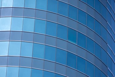 Modern bir binanın birçok dikdörtgen pencereli bir bölümü berrak, parlak mavi gökyüzünü yansıtıyor..