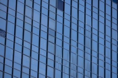 Modern bir binanın birçok dikdörtgen pencereli bir bölümü berrak, parlak mavi gökyüzünü yansıtıyor..