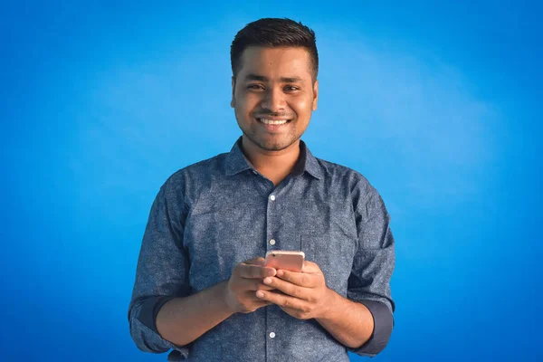 Akıllı telefon tutan ve gülümseyen genç Hintli adamın portresi mavi arka planda izole edilmiş kameraya bakıyor. Hintli iş adamı ve finans kavramı.