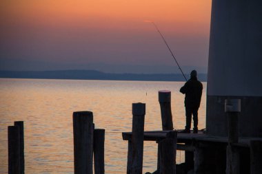 Balık oltasıyla balık tutan balıkçıların silueti, ahşap rıhtım sütunları ve deniz feneri detaylarıyla dolu iskele, gün batımından sonra renkli gökyüzünde deniz suyu üzerinde.