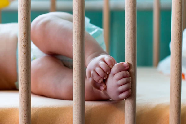 Little feet and cute toes of the 4 months old baby in the baby crib, Baby\'s feet touching wooden pillars of the crib close up, soft blue nude colours