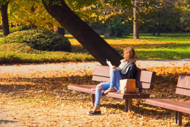Sonbahar güneşli öğleden sonra parkta, genç bayan kitap okuyor, kız elinde alışveriş çantasıyla park bankında oturuyor, sonbahar renkli yapraklar kaldırım ve çimenleri örtüyor, şehir parkında dinleniyor.