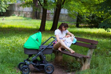Anne ve çocuk, genç anne bebeğini emziriyor ve kucağına alıyor. Bebek arabasıyla parkta, açık hava parkında yürürken duruyor.