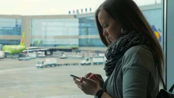 Mujer usando un teléfono inteligente junto a la ventana en el aeropuerto, plano en segundo plano — Vídeos de Stock