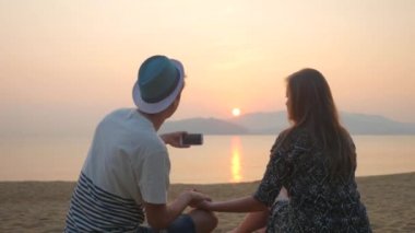 At sunrise beach alarak fotoğraf akıllı telefon ile oturan bir çift. Romantik Çift yaz aylarında Smartphone kullanarak fotoğraf çekmek. Kadın ve erkek tatil günleri tatil seyahat