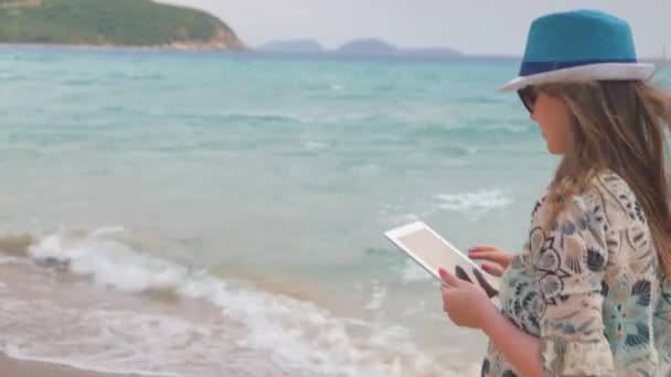 Красива жінка в капелюсі і сонцезахисних окулярах, що йдуть біля моря. Дівчина використовує планшет, стайка-стрілялка — стокове відео