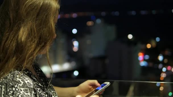 Jolie jeune fille debout sur le balcon et utilisant un téléphone portable à écran tactile. Nuit trouble vue sur la ville avec un magnifique bokeh en arrière-plan . — Video