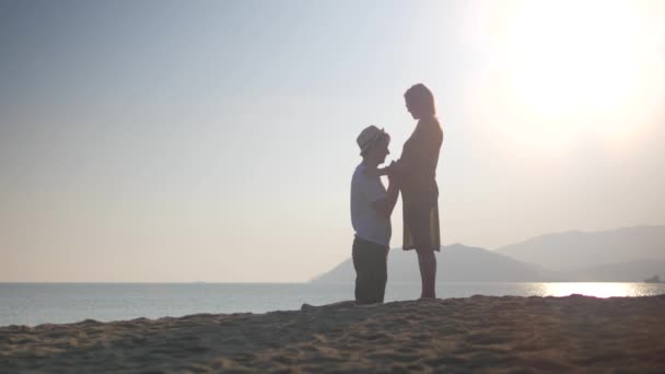 Um casal feliz na praia do pôr-do-sol. Homem de joelhos dobrados acaricia e beija sua barriga esposa — Vídeo de Stock