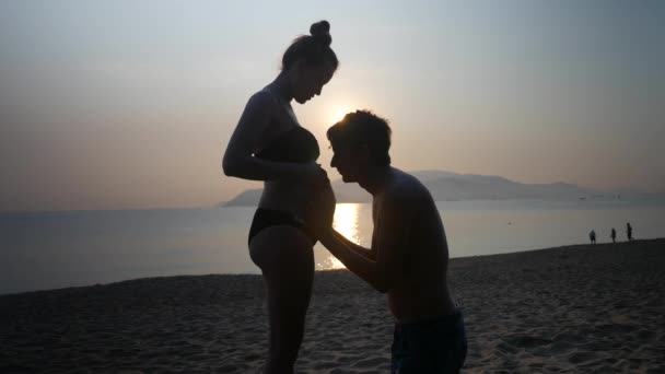 Silhouette di incinta in bikini e romantico uomo in ginocchio piegato accarezzare e baciare la sua pancia mogli su uno sfondo tramonto mare . — Video Stock