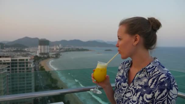 Atractiva joven bebiendo jugo y sonriendo a la cámara mientras mira la vista de la ciudad desde el balcón, Nha Trang, Vietnam . — Vídeos de Stock