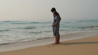 Yalınayak hamile ve kocası bir kum üzerinde duran, birbirlerine sarılma ve muhteşem okyanus manzarası seyir.