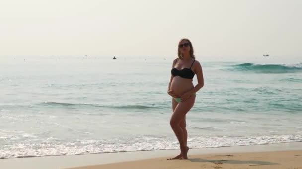 Belle femme enceinte aux cheveux longs en maillot de bain debout sur la plage avec un splendide paysage marin comme arrière-plan . — Video