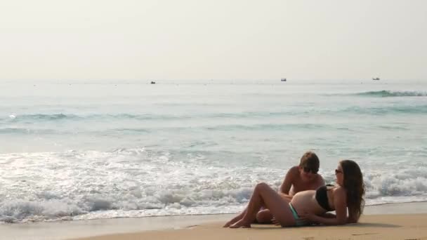 Mujer embarazada y hombre acostado en una playa de arena cerca del agua del mar. Olas salpicando alrededor de ellos . — Vídeo de stock