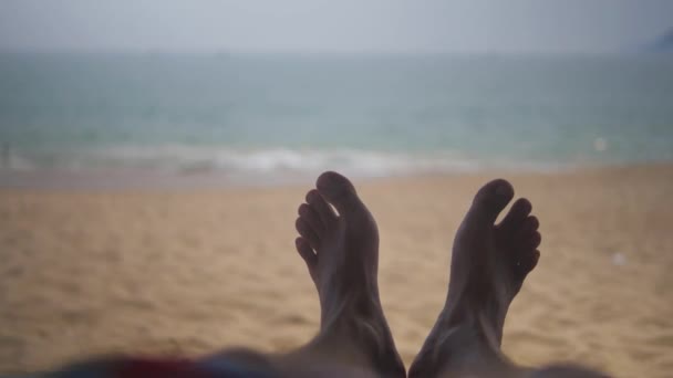 Bare feet of a guy lying on sand with ocean in the background. First-person view — Stock Video