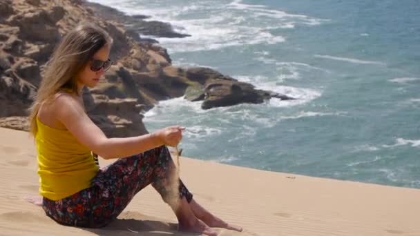 Jeune femme assise sur le sable et jouer avec le sable avec des roches océaniques de la mer sur fond — Video