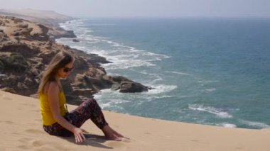 Kum üzerine oturan ve sandfrom el deniz okyanus taşlarla arka plan üzerine düşen genç kadın